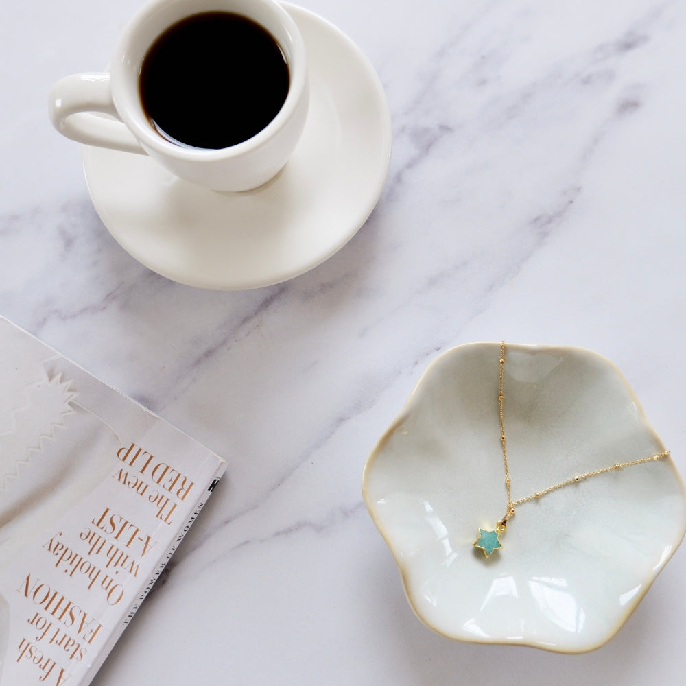 Cup of coffee on a marble surface with a gold amazonite star necklace on a dish and a magazine next to it.