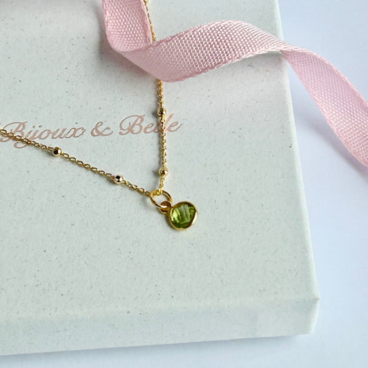 Gold necklace with a vibrant green peridot pendant, resting on a white box with pink ribbon, branded 'Bijoux & Belle'.