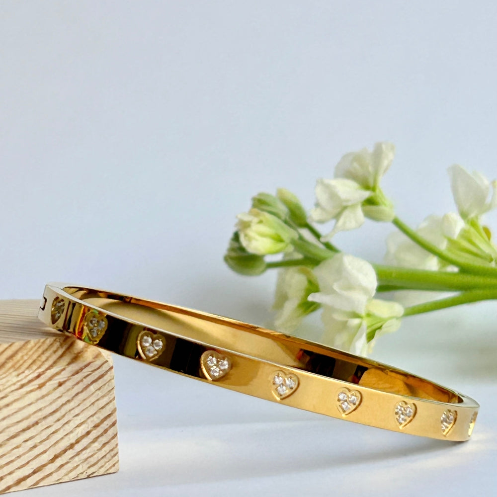 A PVD gold pated bangle with sparkly heart design resting on a wooden block in front of wite flowers.