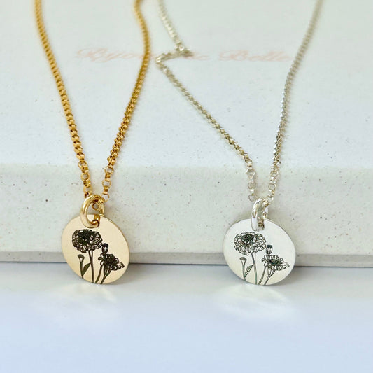 One gold necklace and one silver necklace with circular pendants engraved with the October birth flower (marigold) resting on a white jewellery box.