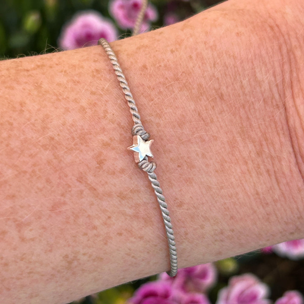 A grey silk friendship bracelet with a star charm on a wrist against a blurred floral background
