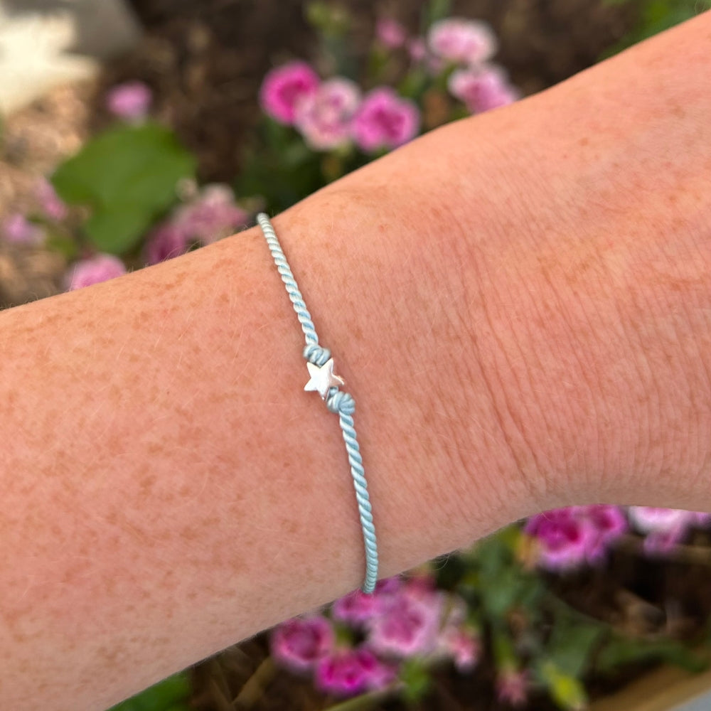 Wrist wearing a light aqua blue silk bracelet with a star charm against a blurred floral background