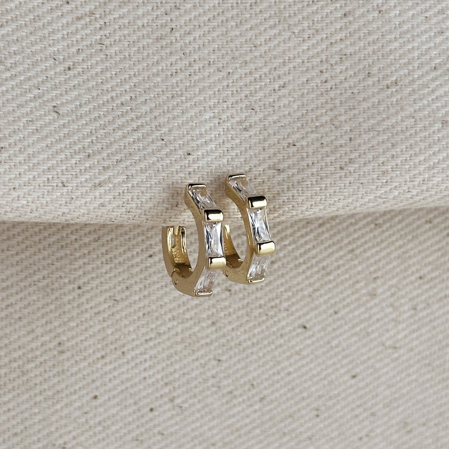 Gold hoop earrings with clear baguette cut cubic zirconia stones on a textured beige background