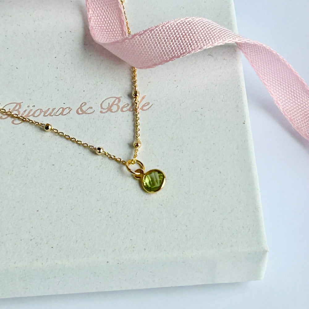 Gold necklace with a vibrant green peridot pendant, resting on a white box with pink ribbon, branded 'Bijoux & Belle'.