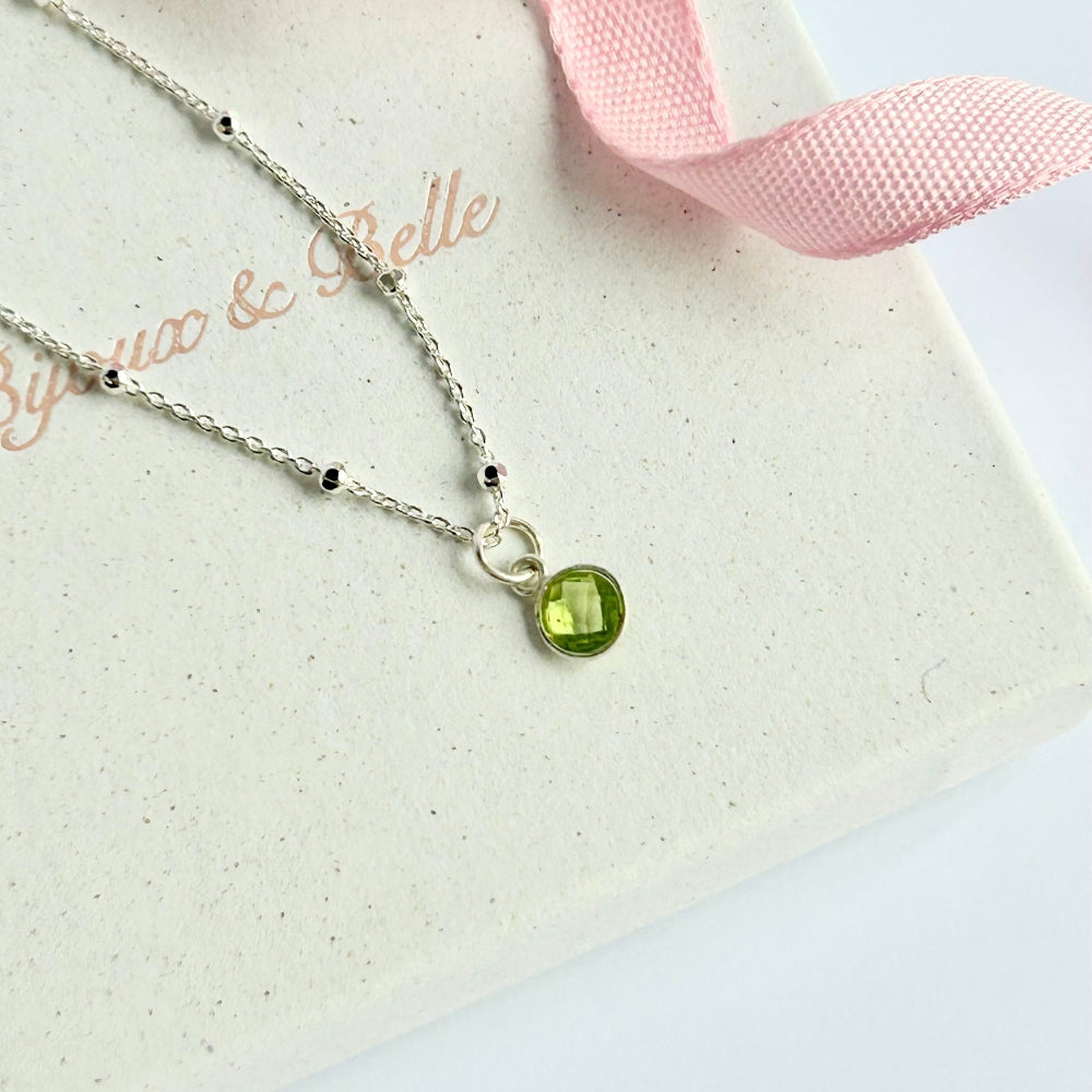 Silver necklace with a vibrant green peridot pendant, resting on a white box with pink ribbon, branded 'Bijoux & Belle'.