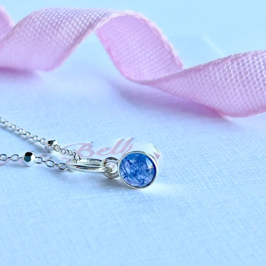Silver necklace with a blueish purple tanzanite pendant , resting on a textured whoie box with pink ribbon.