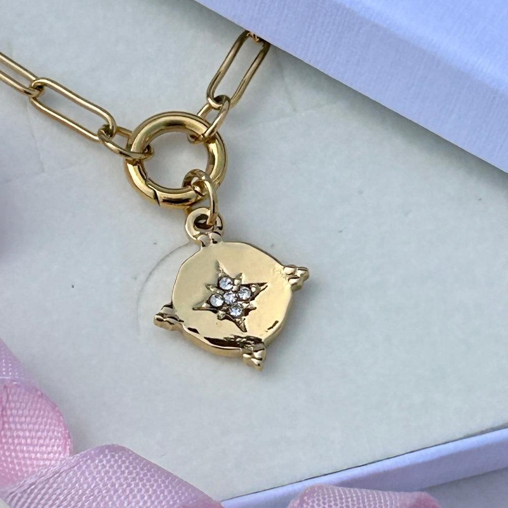 A gold-toned paperclip chain necklace with a small round charm featuring a starburst design set with clear rhinestones, displayed on a light cream jewelry box with pink ribbon accents.