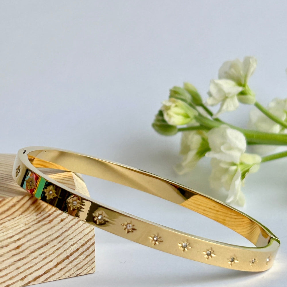 A gold PVD mplated bangle with engraved sparkly star detail resting on a wooden block with white flowers in the background