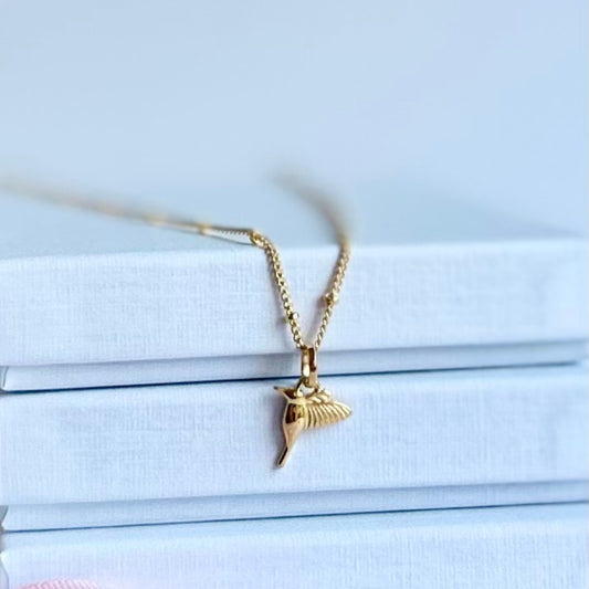 Gold filled hummingbird pendant on a satellite chain, resting on textured white boxes.
