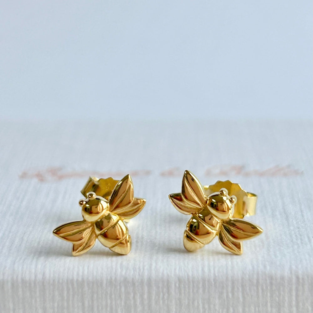 Gold bee-shaped stud earrings resting on a white textured jewellery box.
