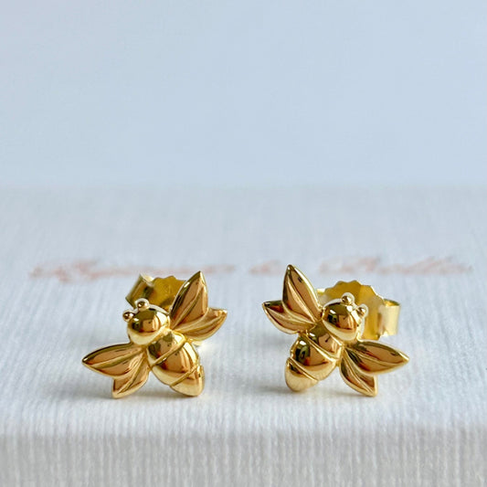 Gold bee-shaped stud earrings resting on a white textured jewellery box.