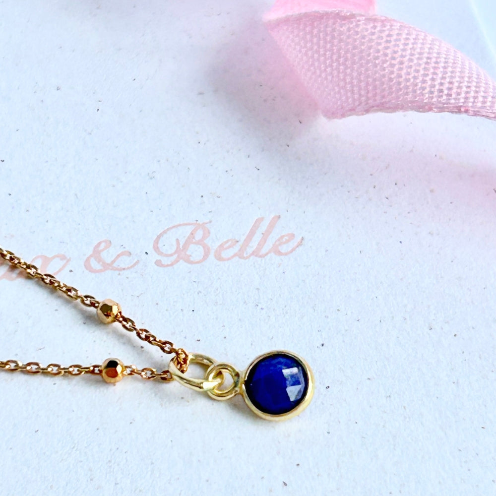 Blue sapphire pendant on a gold satellite chain, resting on a white box with a pink ribbon.