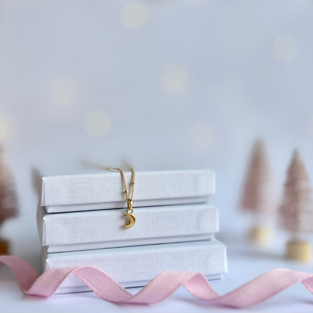Gold necklace with a moon charm on a stack of white boxes with a pink ribbon, blurred decorative elements in the background.