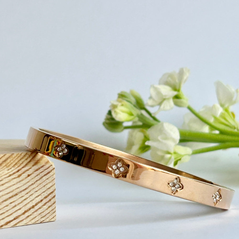 A rose gold PVD plated bangle with a sparkly four leaf clover design resting on a wooden block with white flowers