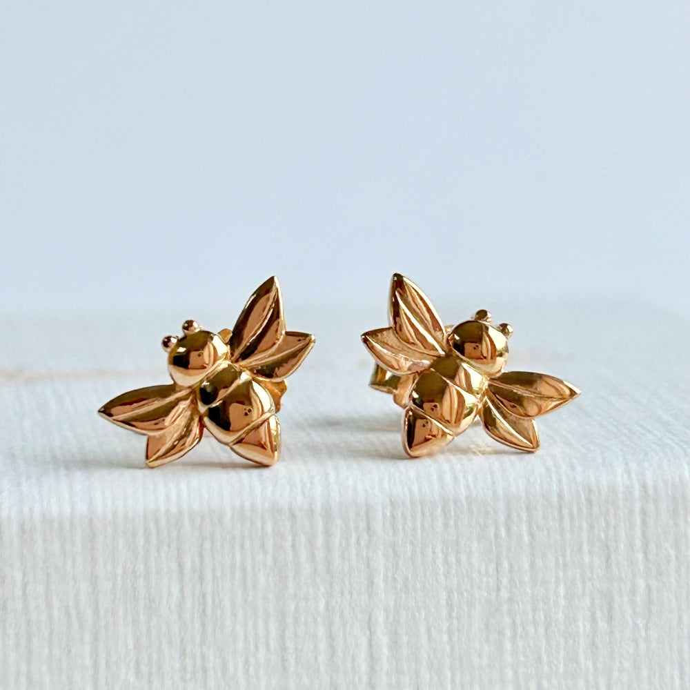 A pair of rose gold bee shaped stud earrings resting on a textured white jewellery box.