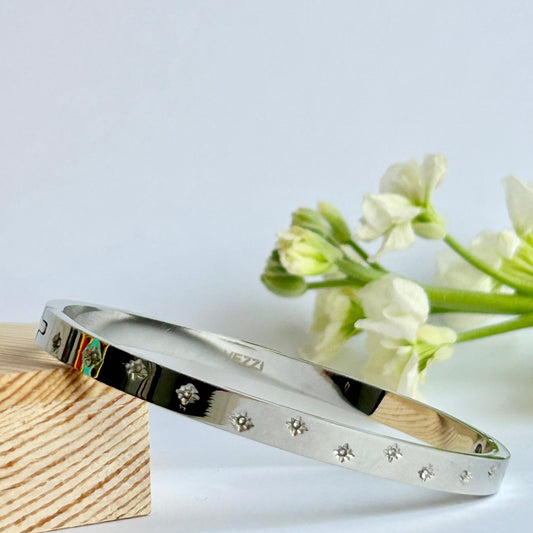 A silver colour bangle with engraved sparkly star design resting on a wooden block with white flowers in the background