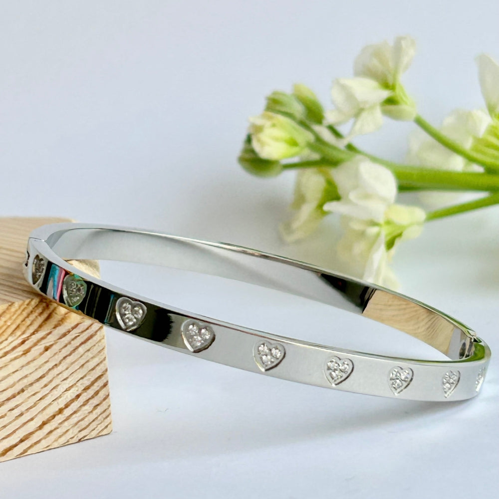 A silver colour bangle with sparkly heart detail on a light background with white flowers behind
