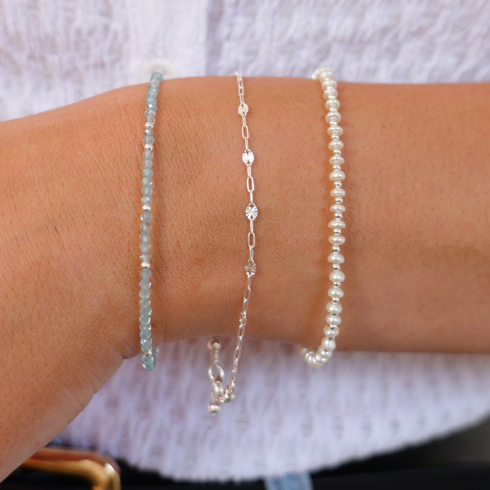 A stack of three bracelets on a wrist against a white top - Apatite tiny gemstone bracelet. sterling silver starburst bracelet and sterling silver button pearl bracelet
