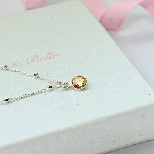 A sterling silver satellite chain with a a warm yellow citrine pendant, resting on a jewellery box branded with 'Bijoux & Belle'.