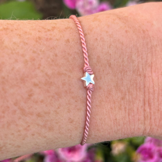 Pink silk bracelet with a star charm on a wrist against a blurred floral background